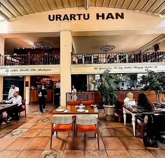 Urartu Han salonunda Urartu Han yazısının altında üst kat balkonu, ahşap masalar, turuncu sandalyeler ve saksı bitkileri.