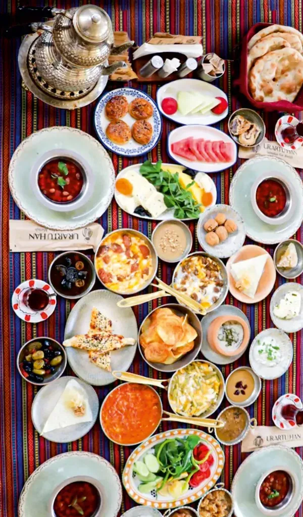 Üstten çekilmiş Van kahvaltısı sofrası: peynirler, menemen, sucuklu yumurta, zeytin, salata, meyve, ekmek ve çay. Görseldeki servis dört kişiliktir; görsel temsilîdir.