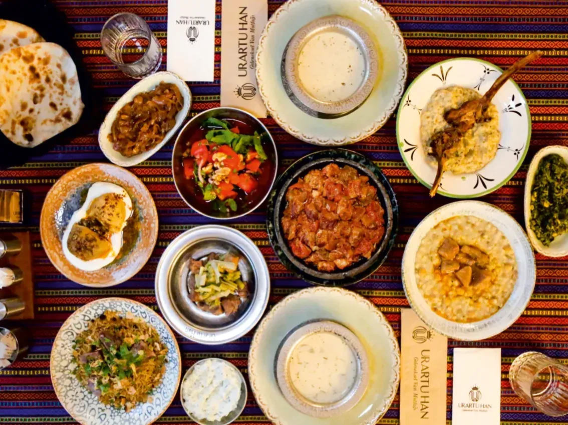 Üstten çekilmiş Han Sofra: iki kâse çorba, salata, mezeler, ekmek ve Van güveci, keledoş, kuzu pirzola gibi yöresel tabaklar; yanlarında Urartu Han logolu peçeteler.