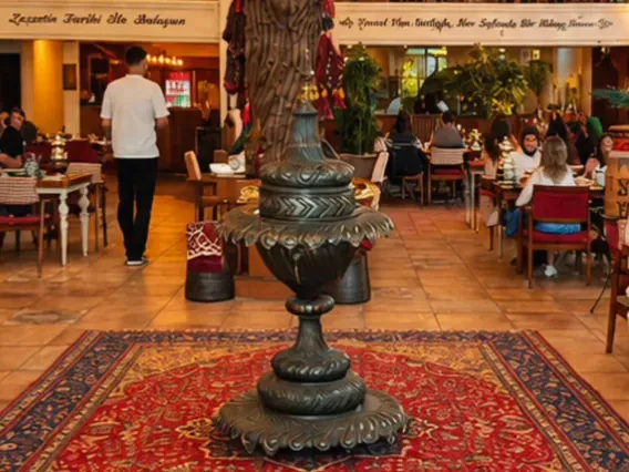 Urartu Han ana salonunda kırmızı desenli halı üzerindeki büyük dekoratif metal vazo; arkada ağaç biçimli dekor ve masalar.