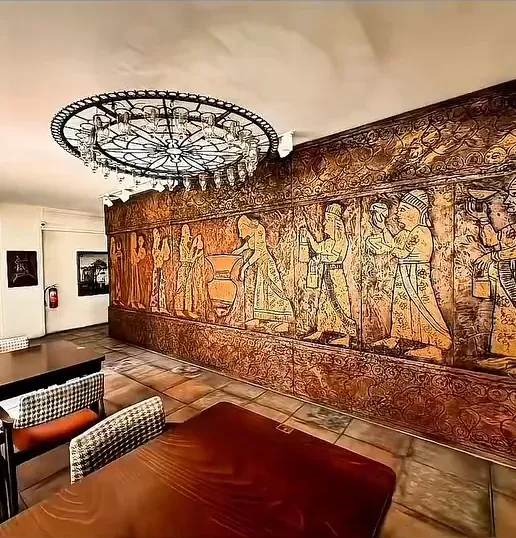 Urartu Han’da tarihî giysili figürlerle işlenmiş bakır tonlu duvar panosu; önünde ahşap masa ve desenli döşemeli sandalyeler, tavanda yuvarlak metal çerçeveli avize.